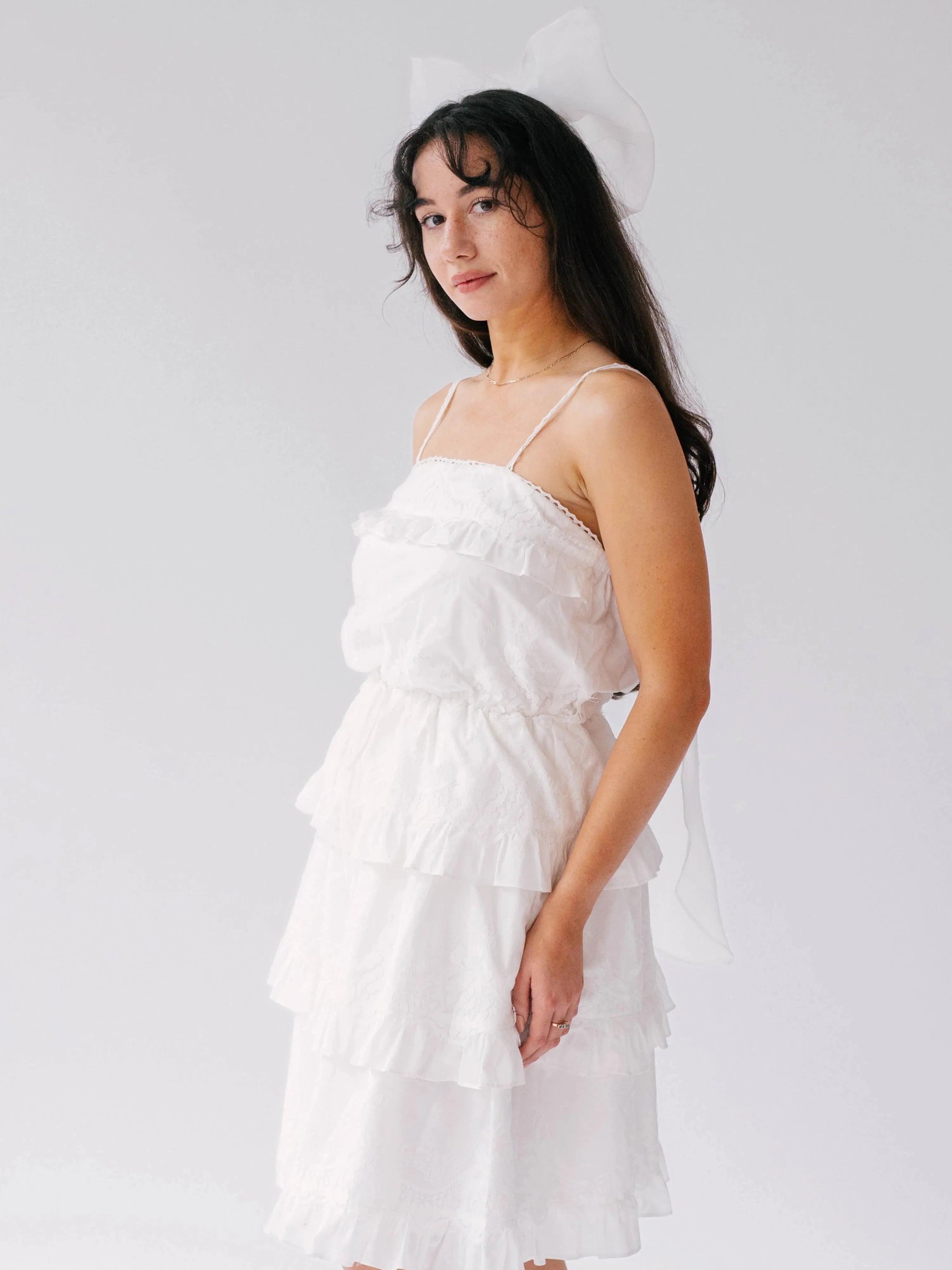 A woman wearing a white dress with a bridal hair bow in her hair, against a plain background.