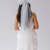 A woman wearing a white ruffle dress with a large white bridal hair bow on her head against a plain background.