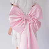 A woman wearing a pink bow belt draped over a white lace dress on a plain background.