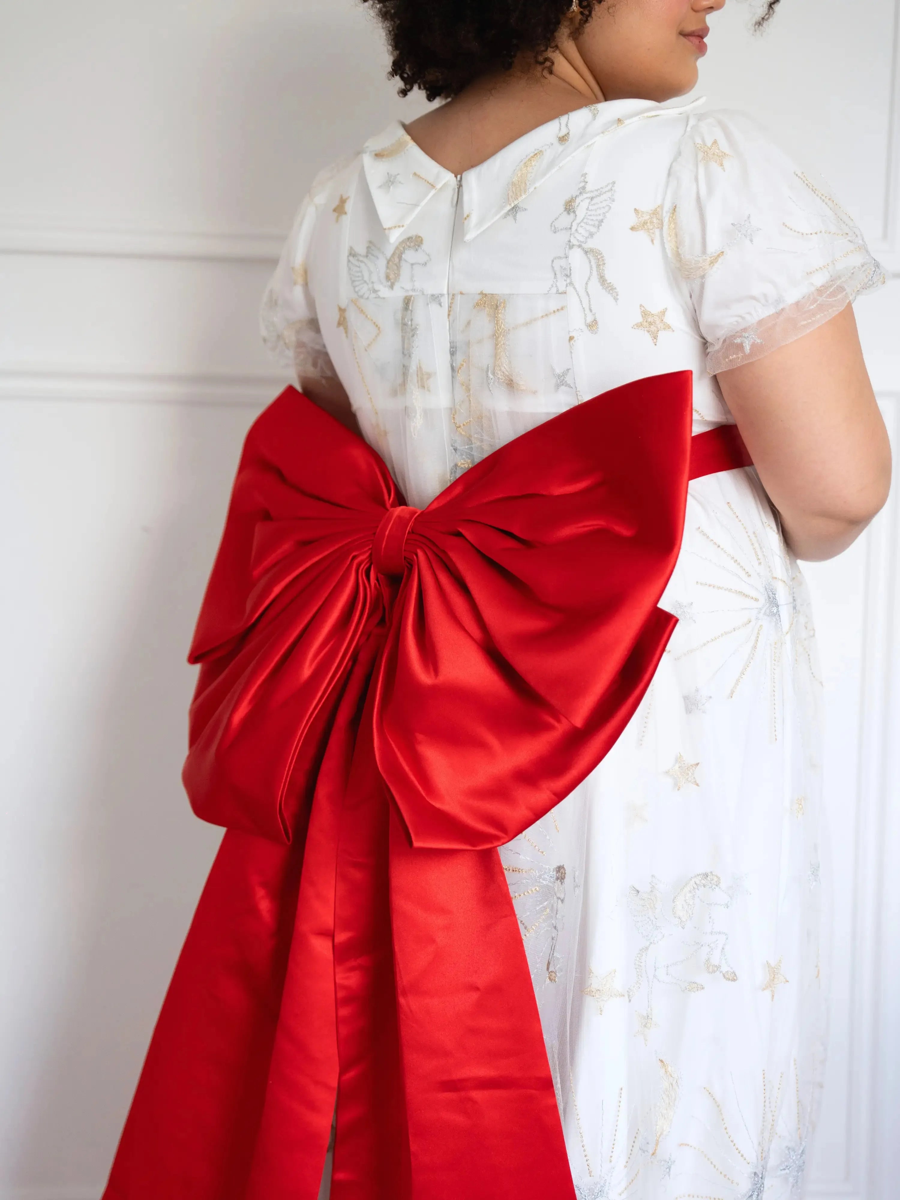 A plus-sized woman wearing the Holiday Red Grand Bow Belt styled on a white dress with gold star patterns.