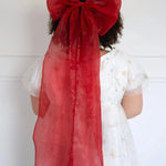 A plus-sized woman wearing a large red hair bow and a lace maxi dress, against a white background.