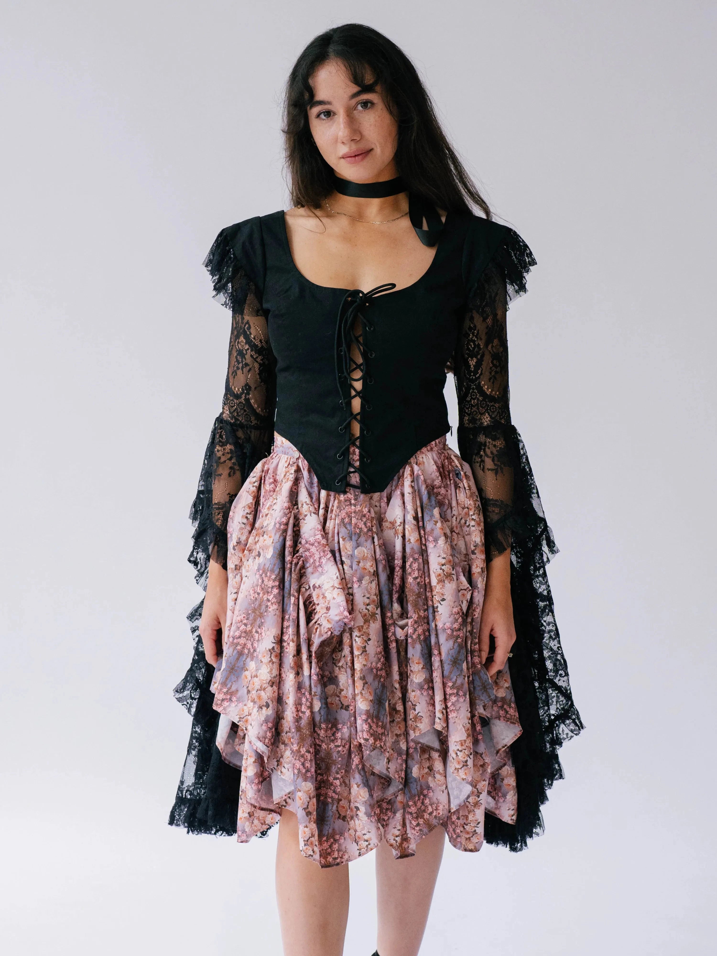 The Guinevere's Revenge Black Corset Top with lace sleeves and front lacing, styled with a floral layered skirt for a romantic look.