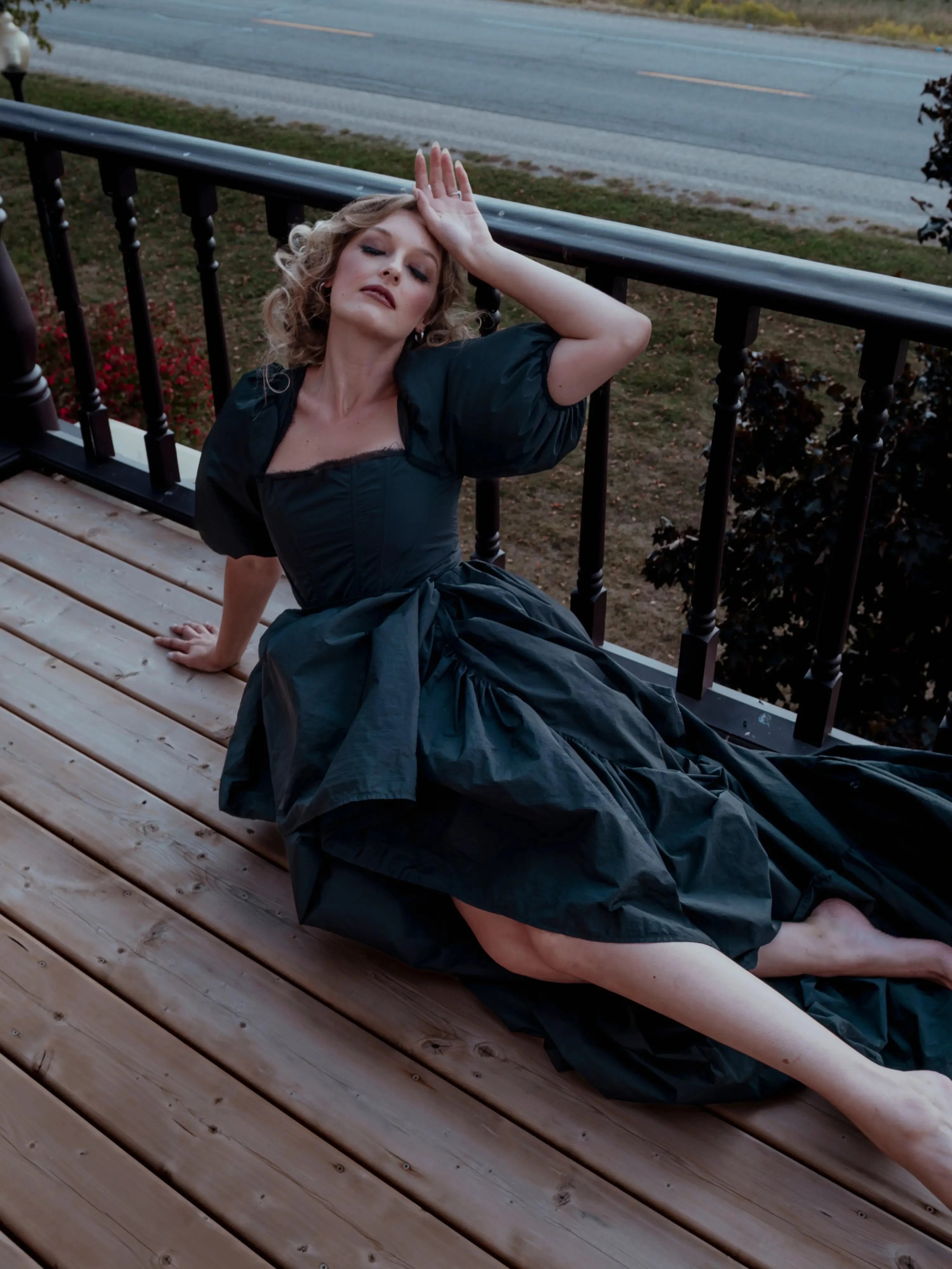 The Josephine Renaissance Gown in deep green with puff sleeves and corset bodice, styled dramatically on a wooden balcony. Gothic fairytale maxi dress.