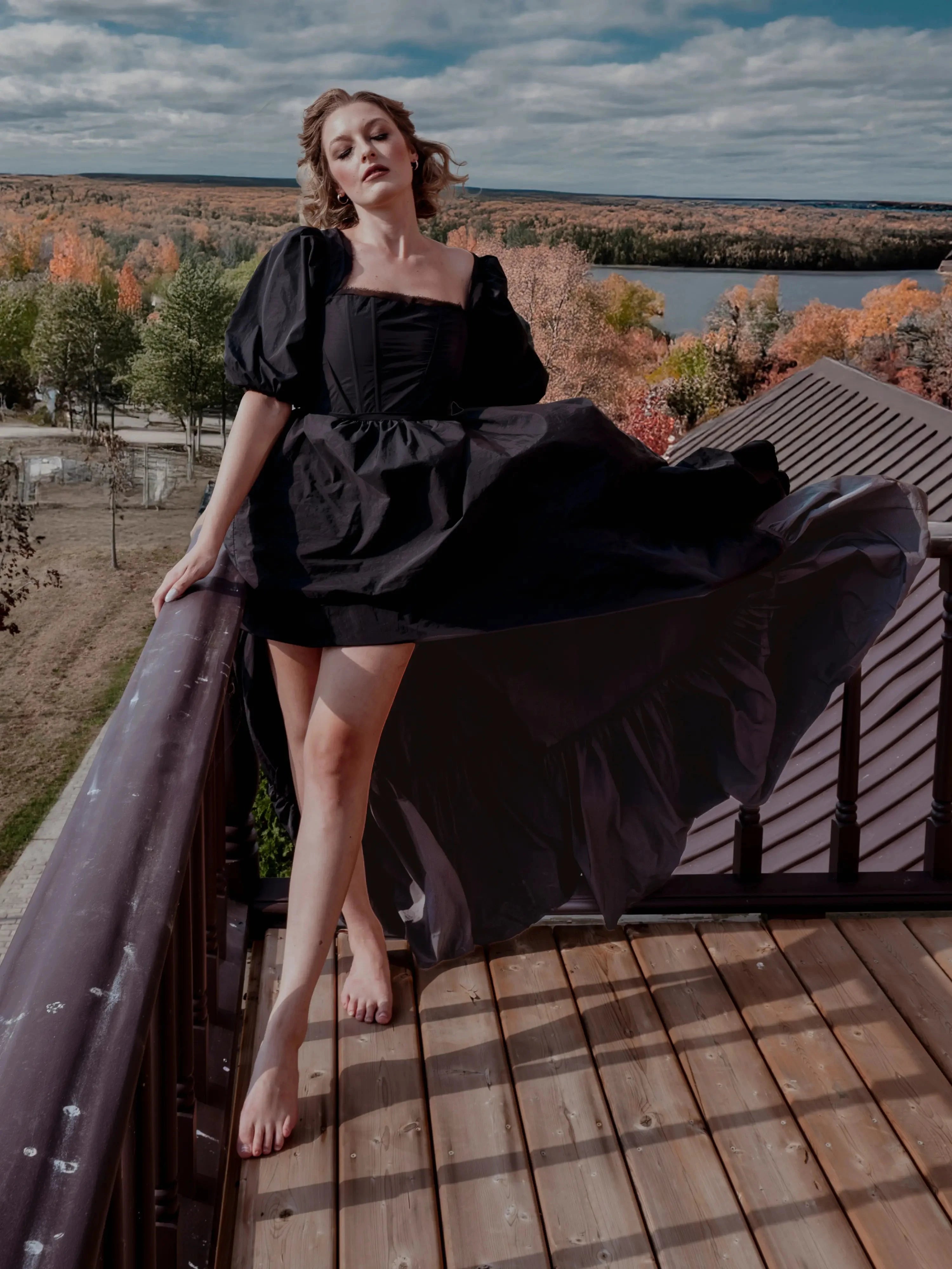 The Josephine Renaissance Gown in black with corset bodice and puff sleeves, flowing dramatically on a balcony with autumn trees in the background.