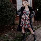 A woman in a floral dress and long black coat is standing in front of a brick building.