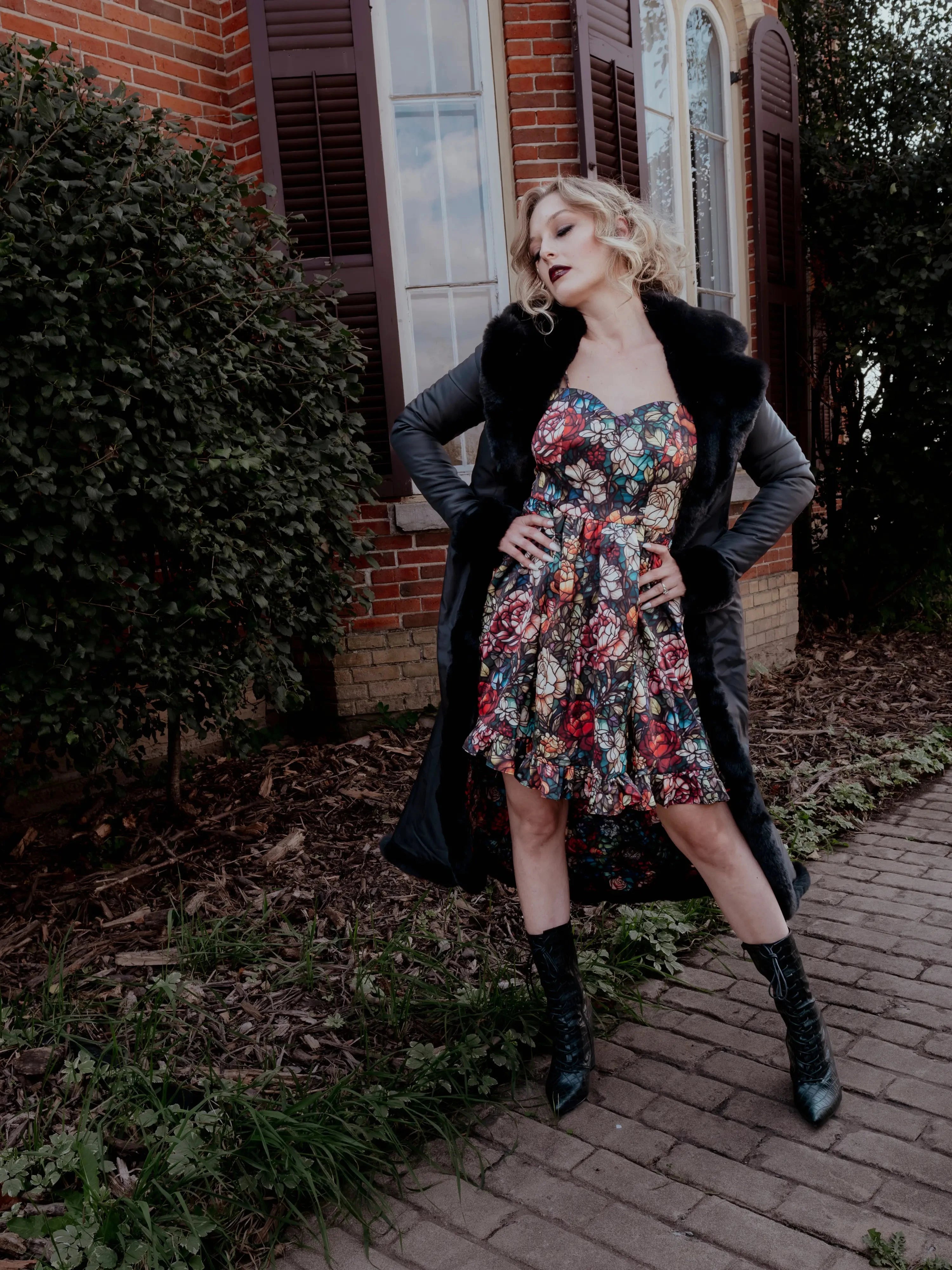 A woman in a floral dress and long black coat is standing in front of a brick building.