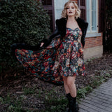 A woman in a floral dress and a long black coat is standing outdoors near a brick building.
