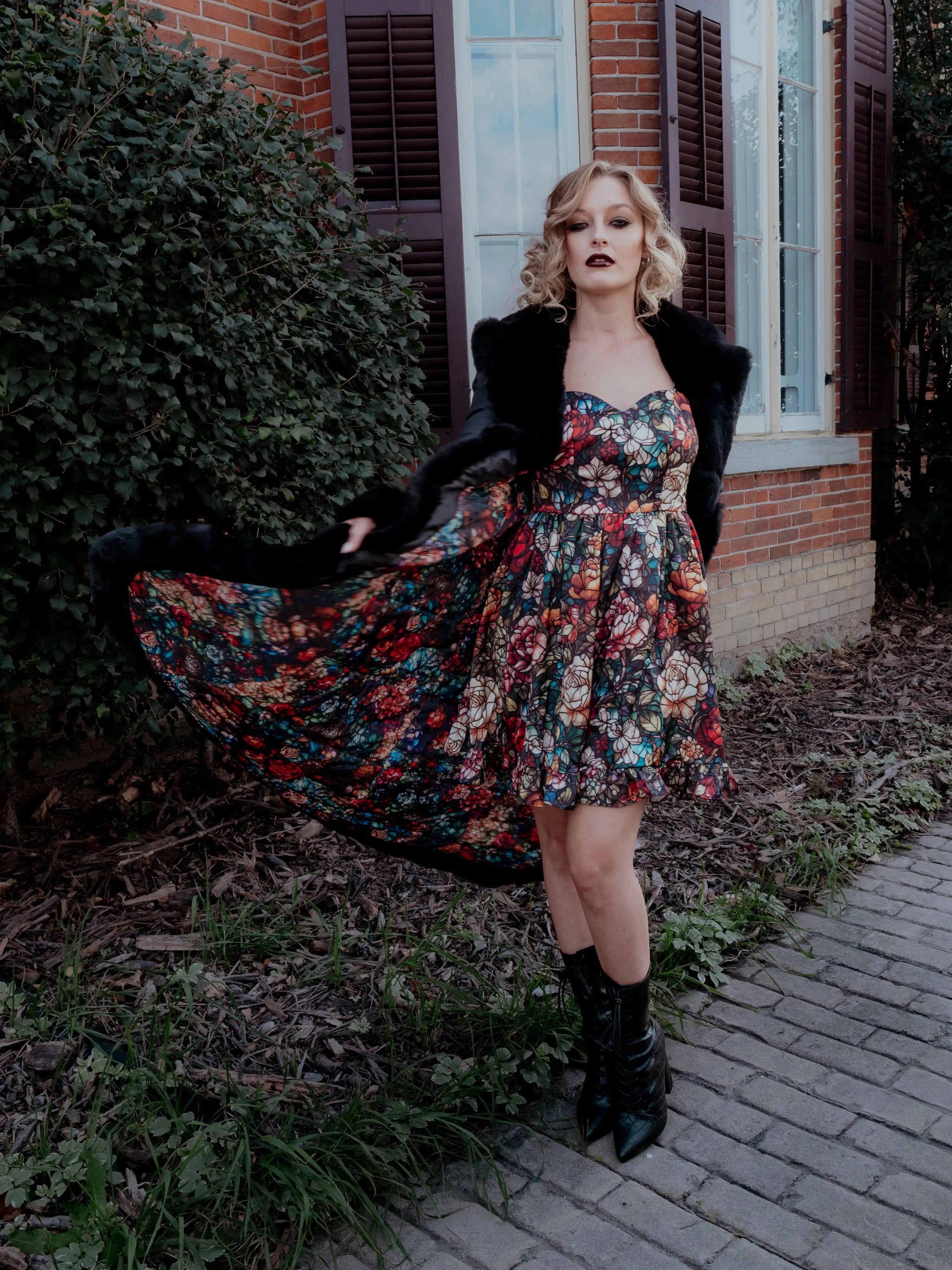 A woman in a floral dress and a long black coat is standing outdoors near a brick building.