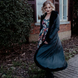 A woman in a long black coat and a floral dress is standing outside near a brick building.