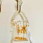 Clear mini bucket bag with gold lock and white ribbon hanging on a hook against a white background.
