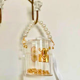 Clear mini bucket bag with gold lock and white ribbon hanging on a hook against a white background.