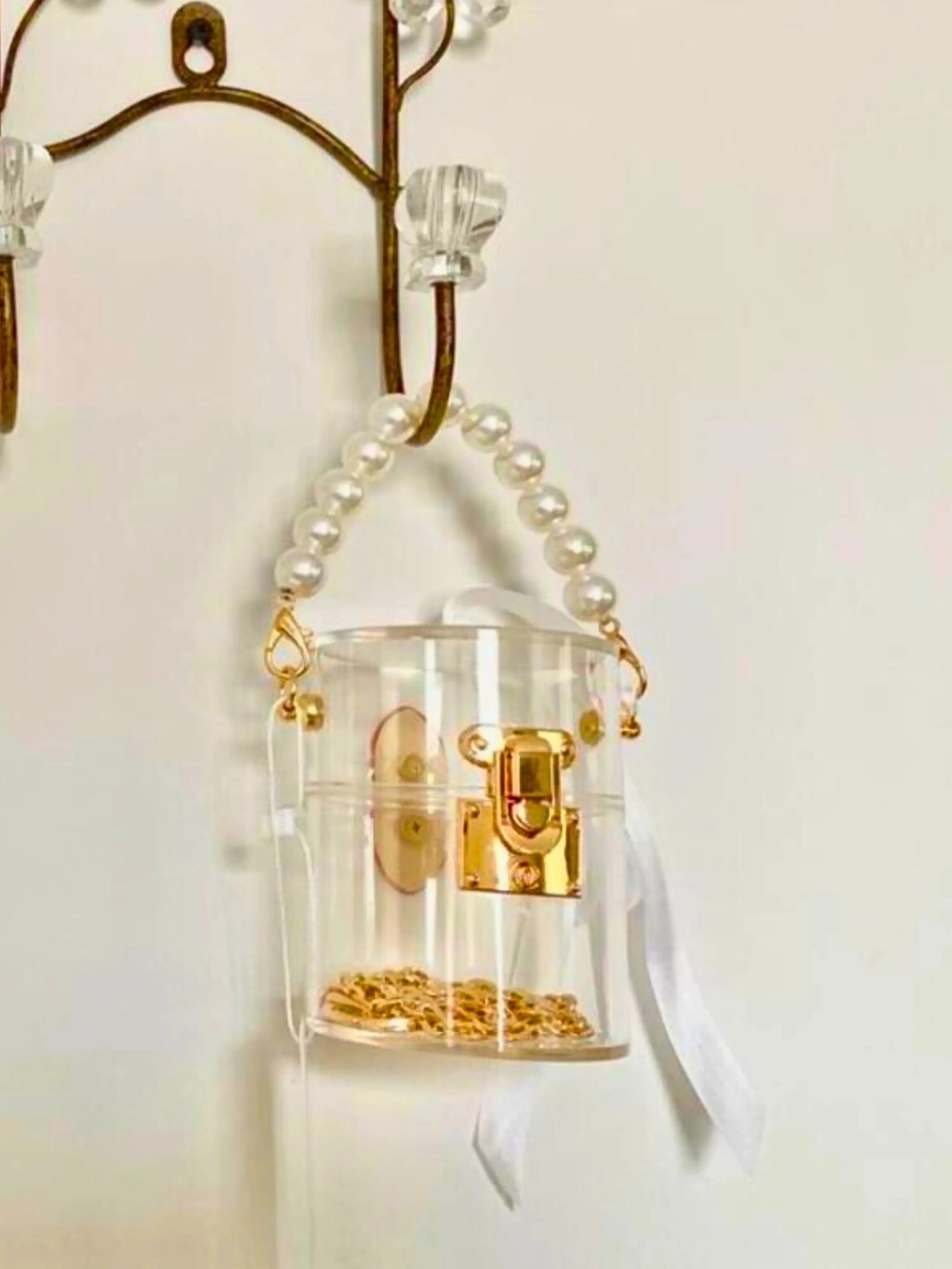 Clear mini bucket bag with gold lock and white ribbon hanging on a hook against a white background.