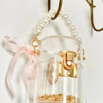 Clear mini bucket bag with gold lock and pink ribbon hanging on a hook against a white background.
