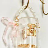 Clear mini bucket bag with gold lock and pink ribbon hanging on a hook against a white background.