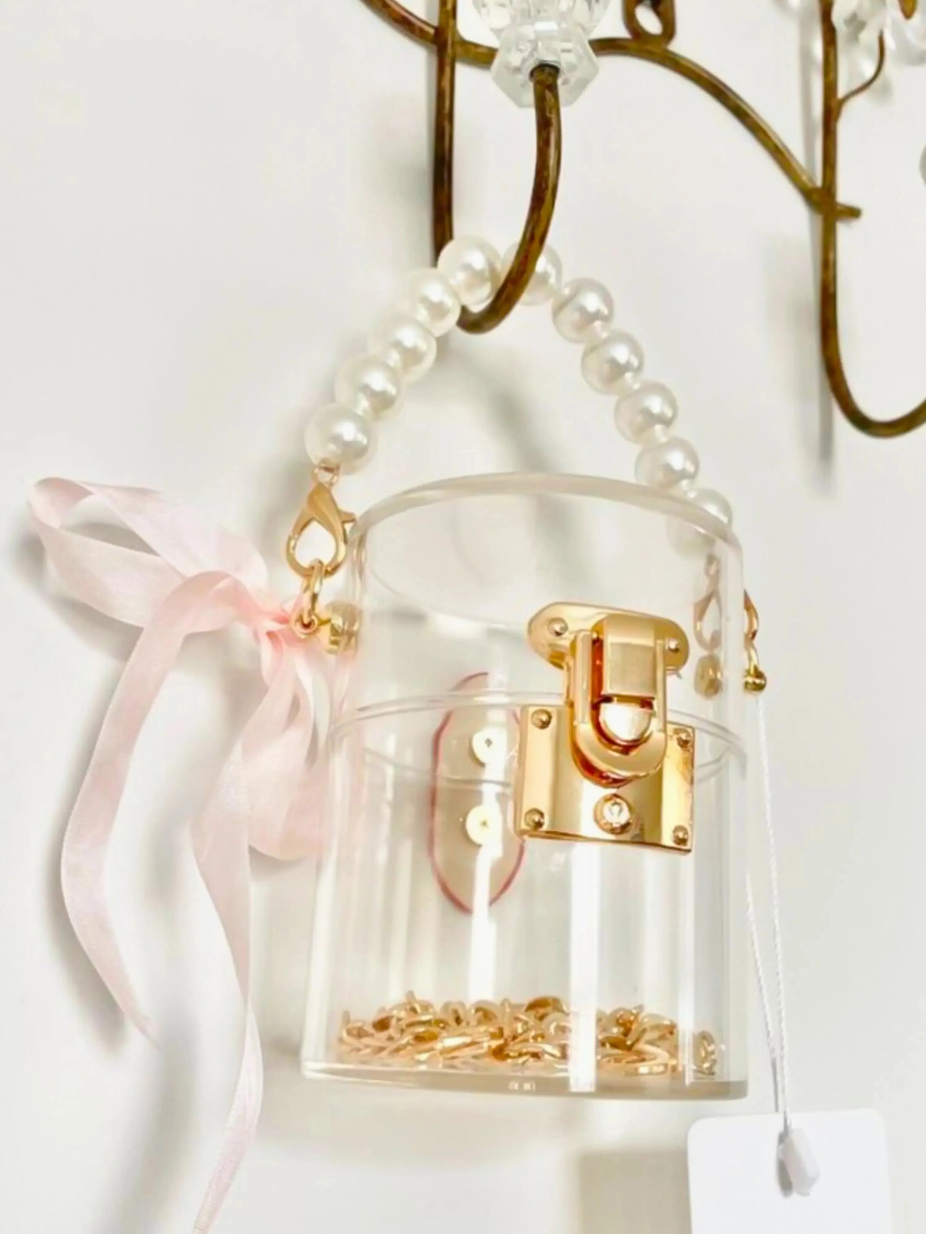 Clear mini bucket bag with gold lock and pink ribbon hanging on a hook against a white background.