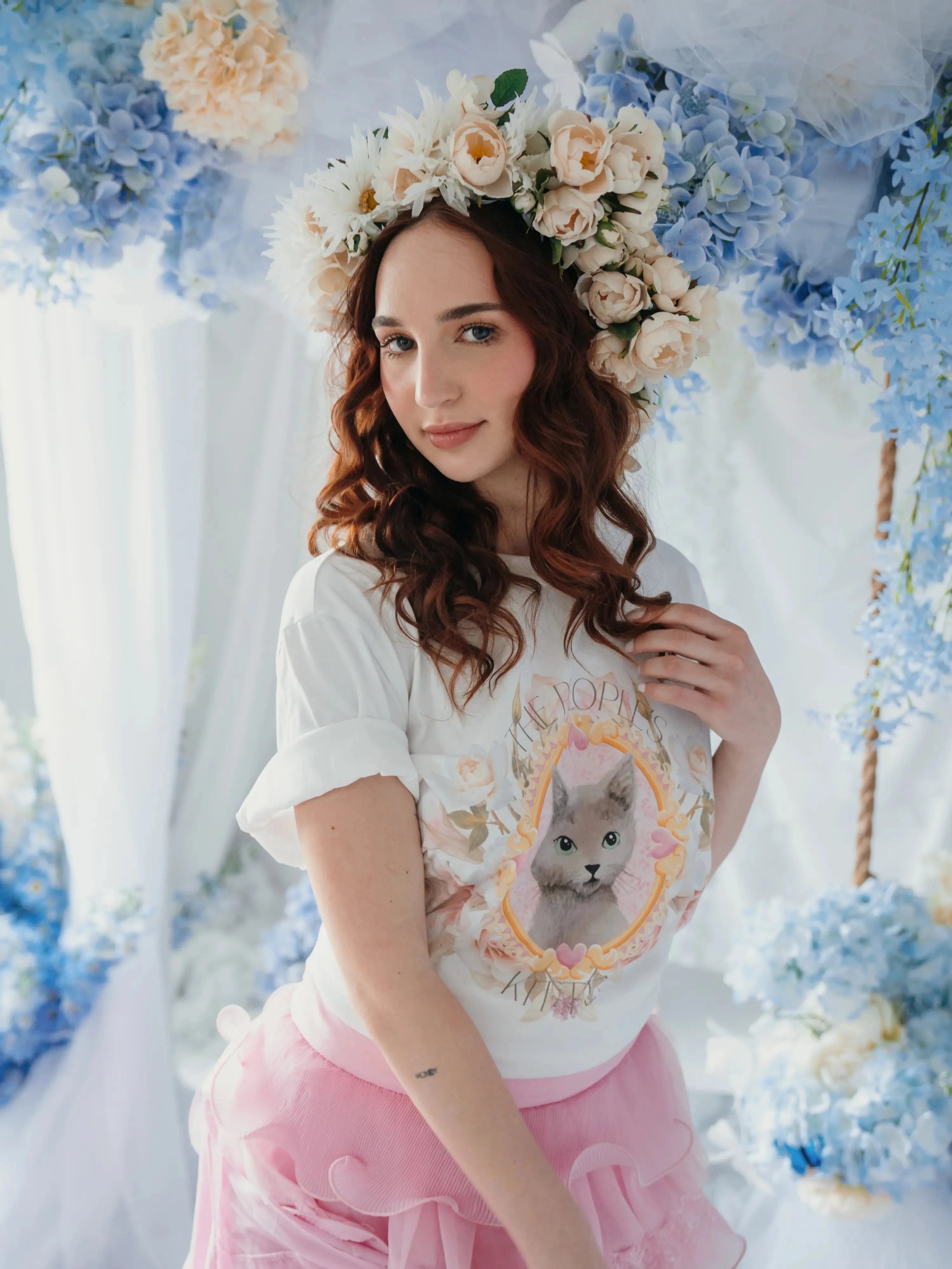 A woman wearing the Princess Kitten Tee styled with a floral crown and pink ruffled skirt, featuring whimsical cat artwork by Kaeti MacNeil.