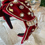 The Little Bit of Blair Red Velvet Headband adds regal charm with its ornate gold details, pearls, and rhinestones on deep red velvet fabric.