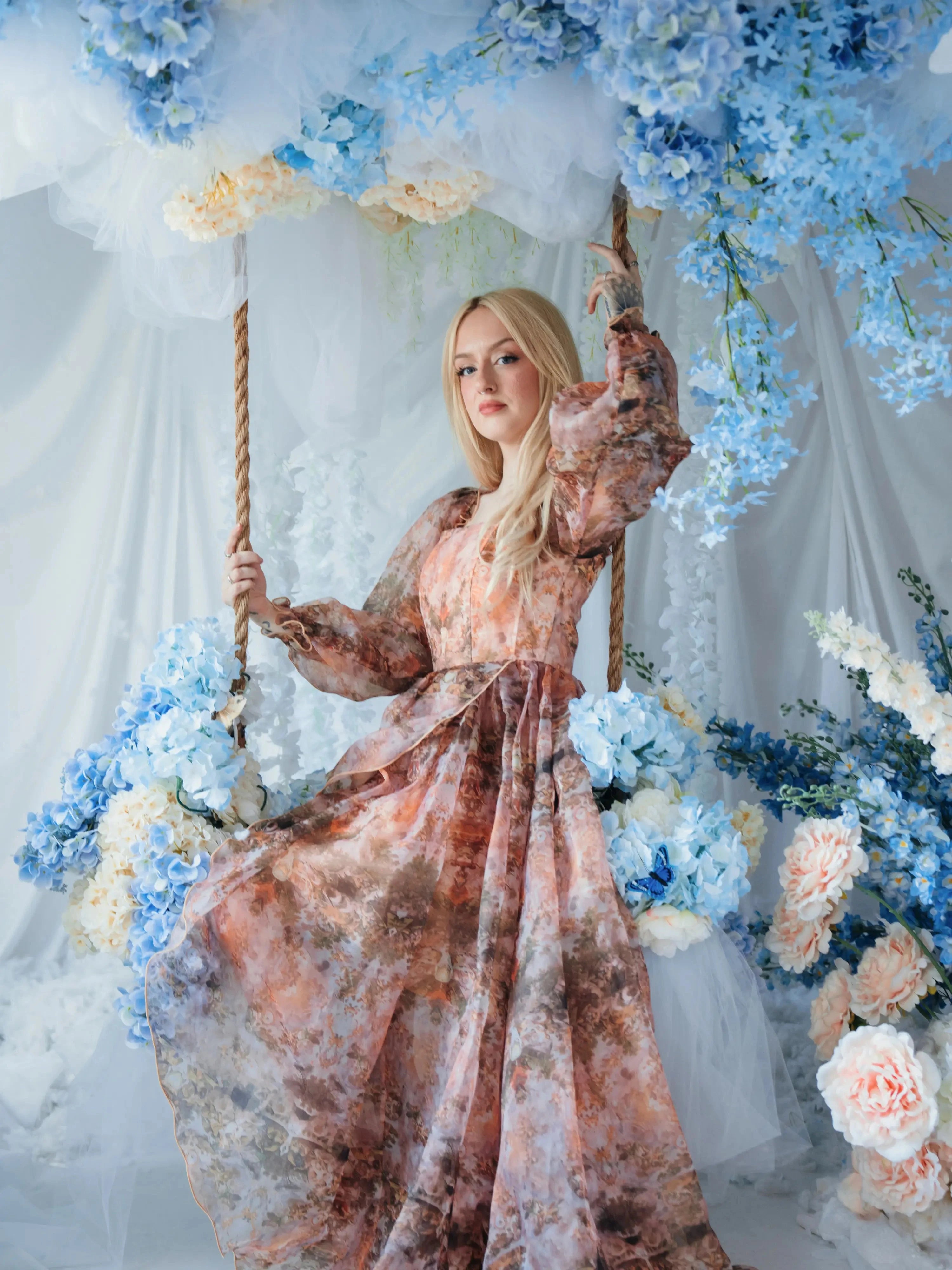 The Ode to Jove Twilight Reverie Ball Gown with baroque fresco print, worn on a floral swing surrounded by blue hydrangeas and white drapery.
