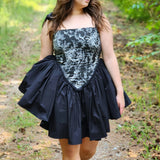 A woman in a black corset mini dress is walking along a scenic path in a forest. A perfect fairytale setting.