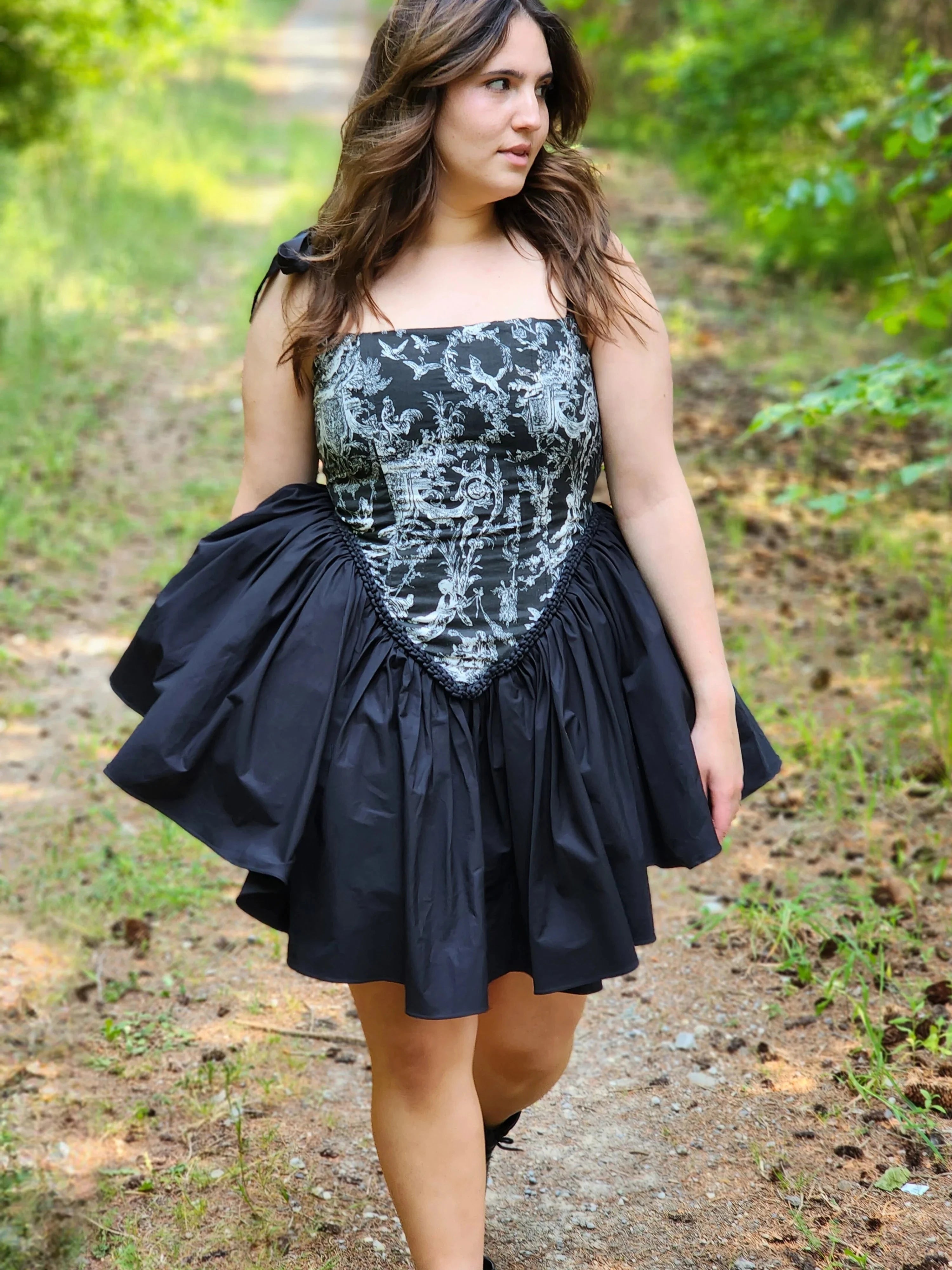 A woman in a black corset mini dress is walking along a scenic path in a forest. A perfect fairytale setting.