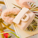 The Petit Agneau Tiny Purse in ivory with faux fur handle and gold clasp, styled on a decorative book for chic micro handbag appeal.