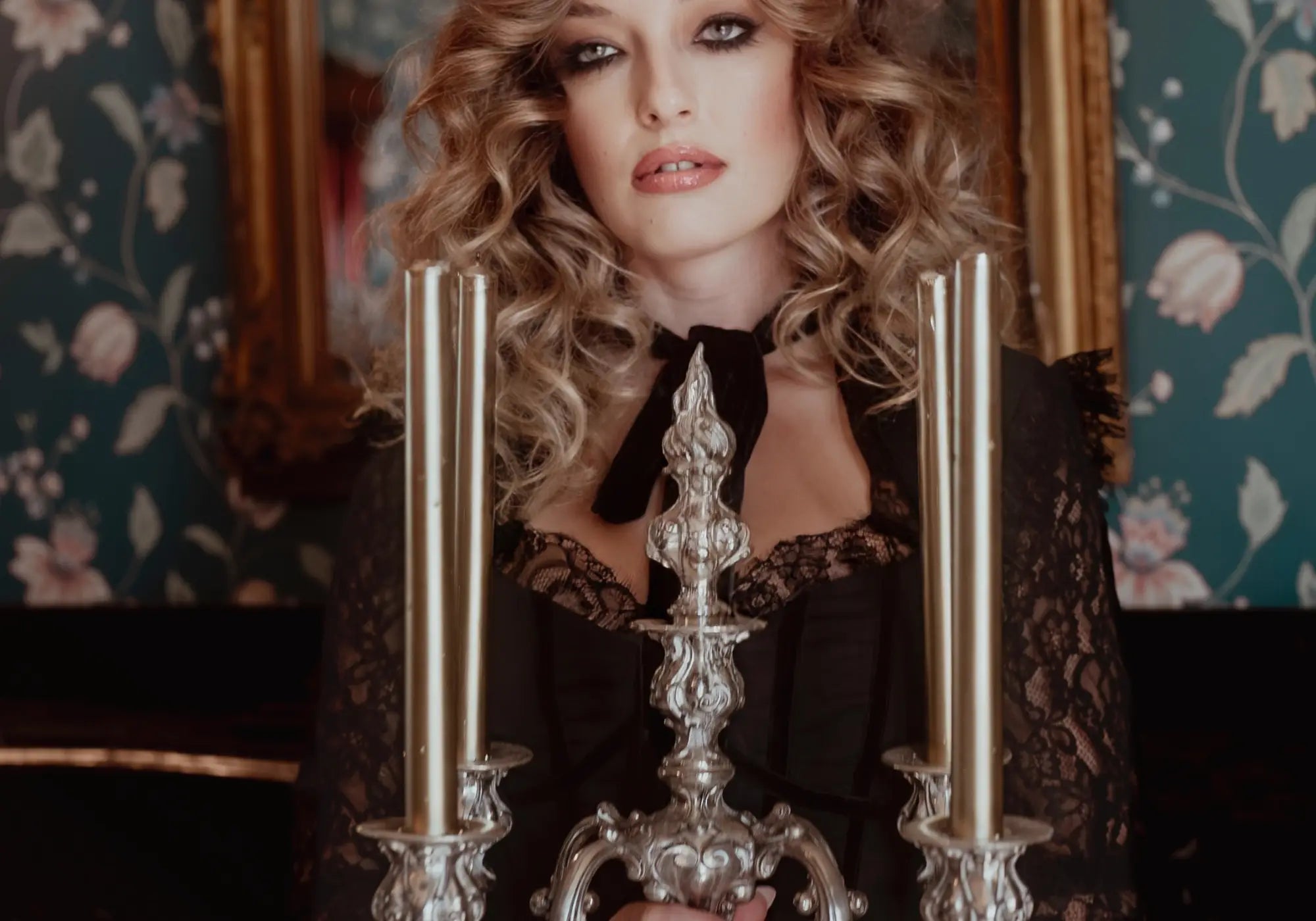 A woman holding a silver candelabrum with candles in an ornate room, wearing a long black lace dress.