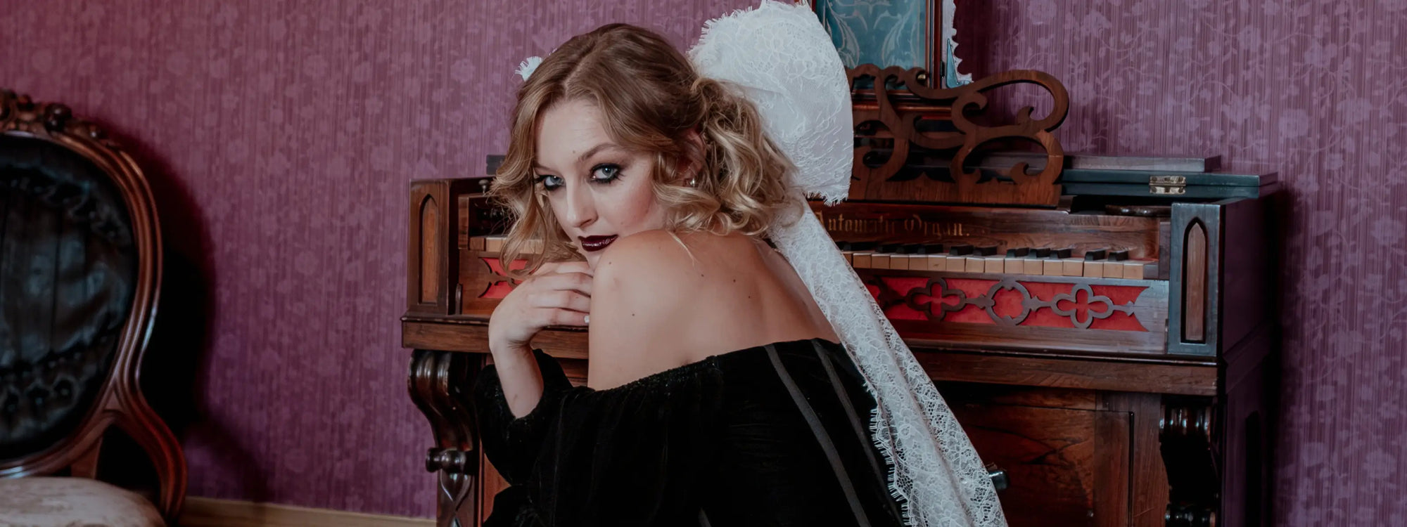 A woman wearing a black velvet mini dress and a white lace hair bow in an antique room with a piano.