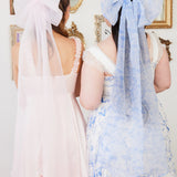 Two dresses with bows. The midi dress on the left is the Ballerina Pink Satin Midi Dress paired with The Ballerina Pink Grand Hair Bow. The corset maxi dress on the right is the Blue Toile Angel Dress paired with The Blue Toile Large Hair Bow.