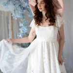 Romantic Belladonna Lace Dress with flutter sleeves and flowing train, styled in a floral wedding setting with pastel blue accents.