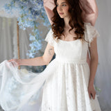Romantic Belladonna Lace Dress with flutter sleeves and flowing train, styled in a floral wedding setting with pastel blue accents.