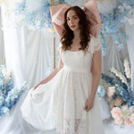 Belladonna Lace Dress styled in dreamy floral backdrop, showcasing lace bodice, soft sleeves, and flowing high-low hemline.