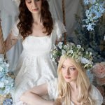 Two models in Belladonna Lace Dresses with floral crowns, seated on a swing surrounded by pastel wedding florals and lace details.