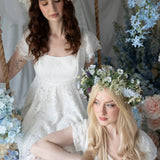 Two models in Belladonna Lace Dresses with floral crowns, seated on a swing surrounded by pastel wedding florals and lace details.