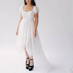 Belladonna Lace Dress midi bridal gown with high-low hem and trailing tulle train, modeled in studio with black platform heels.