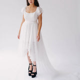 Belladonna Lace Dress midi bridal gown with high-low hem and trailing tulle train, modeled in studio with black platform heels.