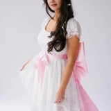 Belladonna Lace Dress styled with pink satin bow sash, highlighting romantic bridal design with lace and high-low silhouette.