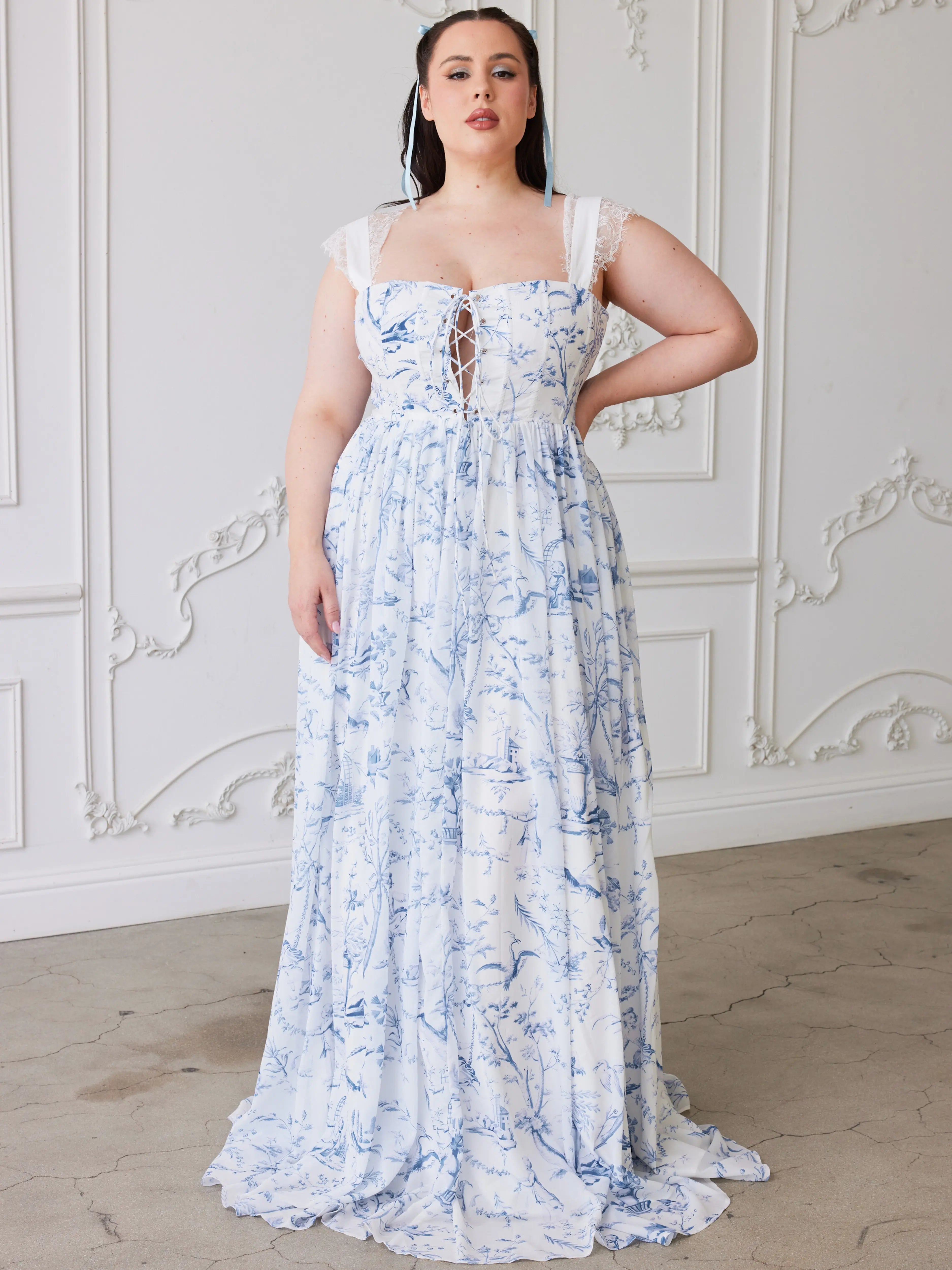 A floor-length white dress with a delicate floral blue toile print. Dress has a corset-style bodice and thin straps with lace details.