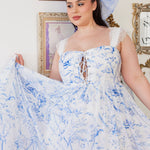 A flowing white midi dress with delicate blue toile patterns and a corset-style bodice. Paired with a large hair bow that has the same pattern.