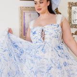 A flowing white midi dress with delicate blue toile patterns and a corset-style bodice. Paired with a large hair bow that has the same pattern.