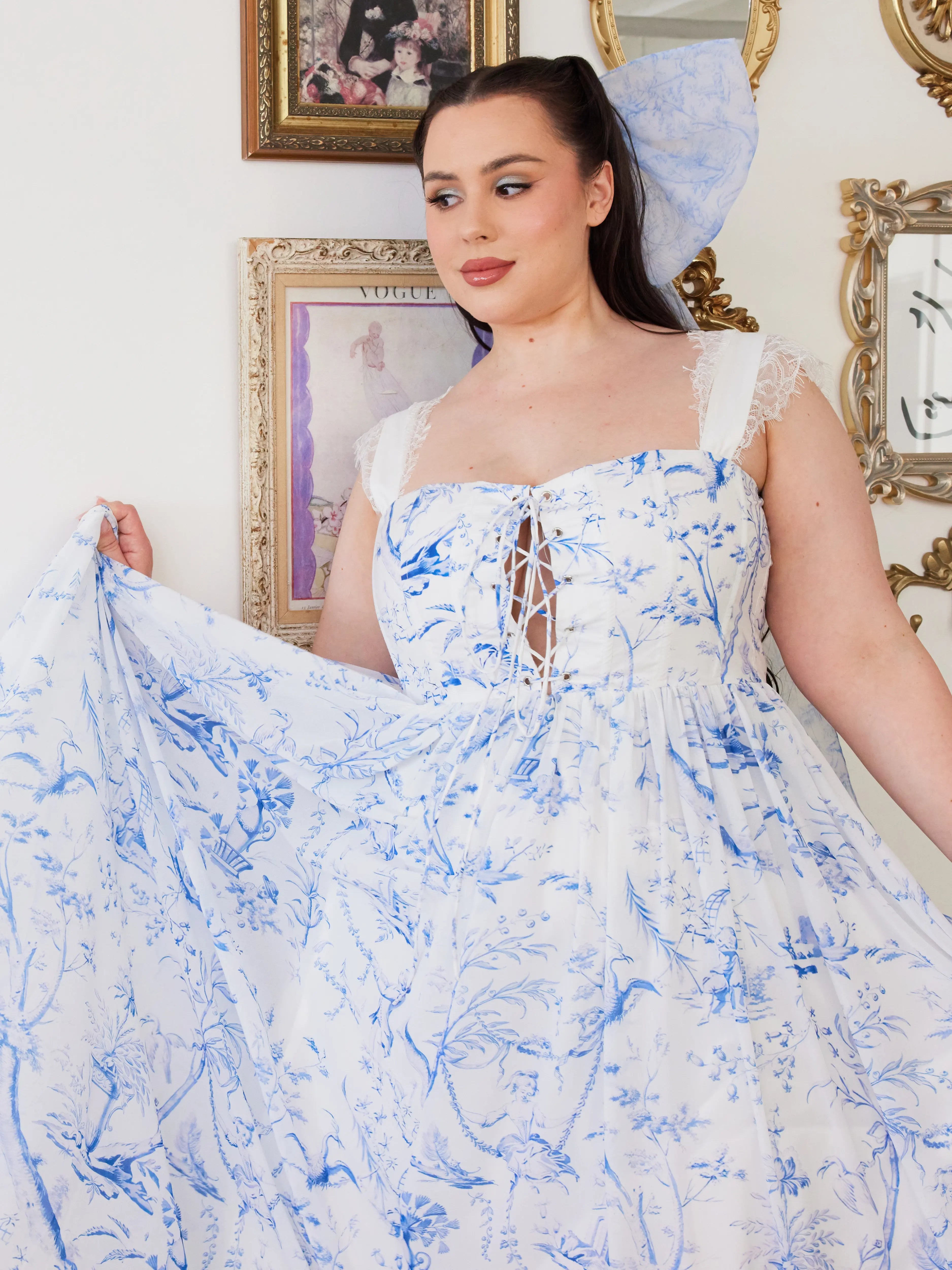 A flowing white midi dress with delicate blue toile patterns and a corset-style bodice. Paired with a large hair bow that has the same pattern.