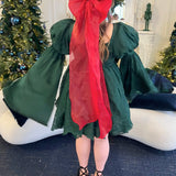 Green mini dress with bell sleeves featuring a giant red Christmas hair bow on a model in a Christmas setting.