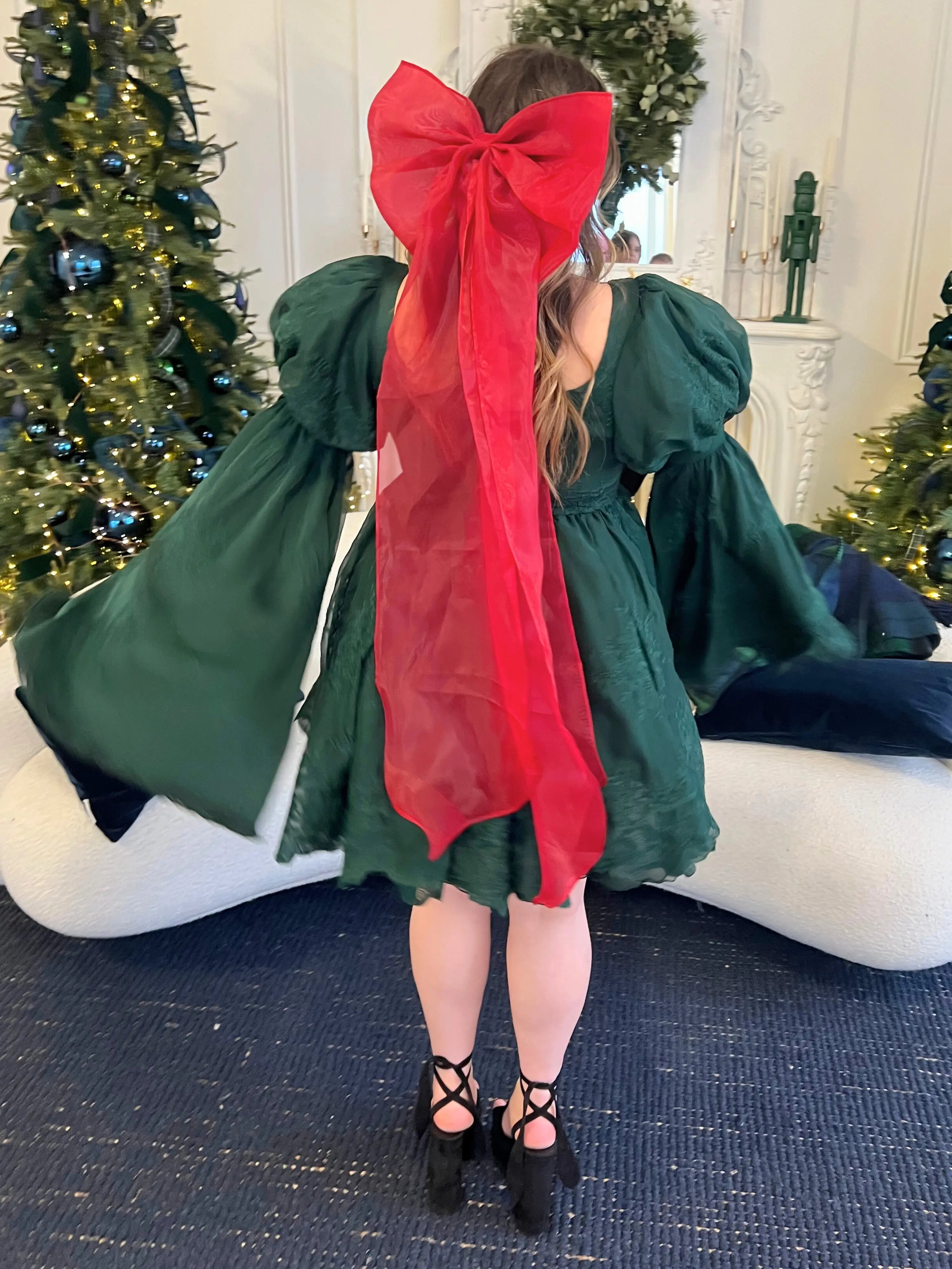 Green mini dress with bell sleeves featuring a giant red Christmas hair bow on a model in a Christmas setting.