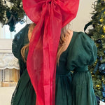 Bright red giant flutter bow on long a model with blonde hair wearing a green holiday mini dress by a Christmas tree.