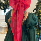 Bright red giant flutter bow on long a model with blonde hair wearing a green holiday mini dress by a Christmas tree.