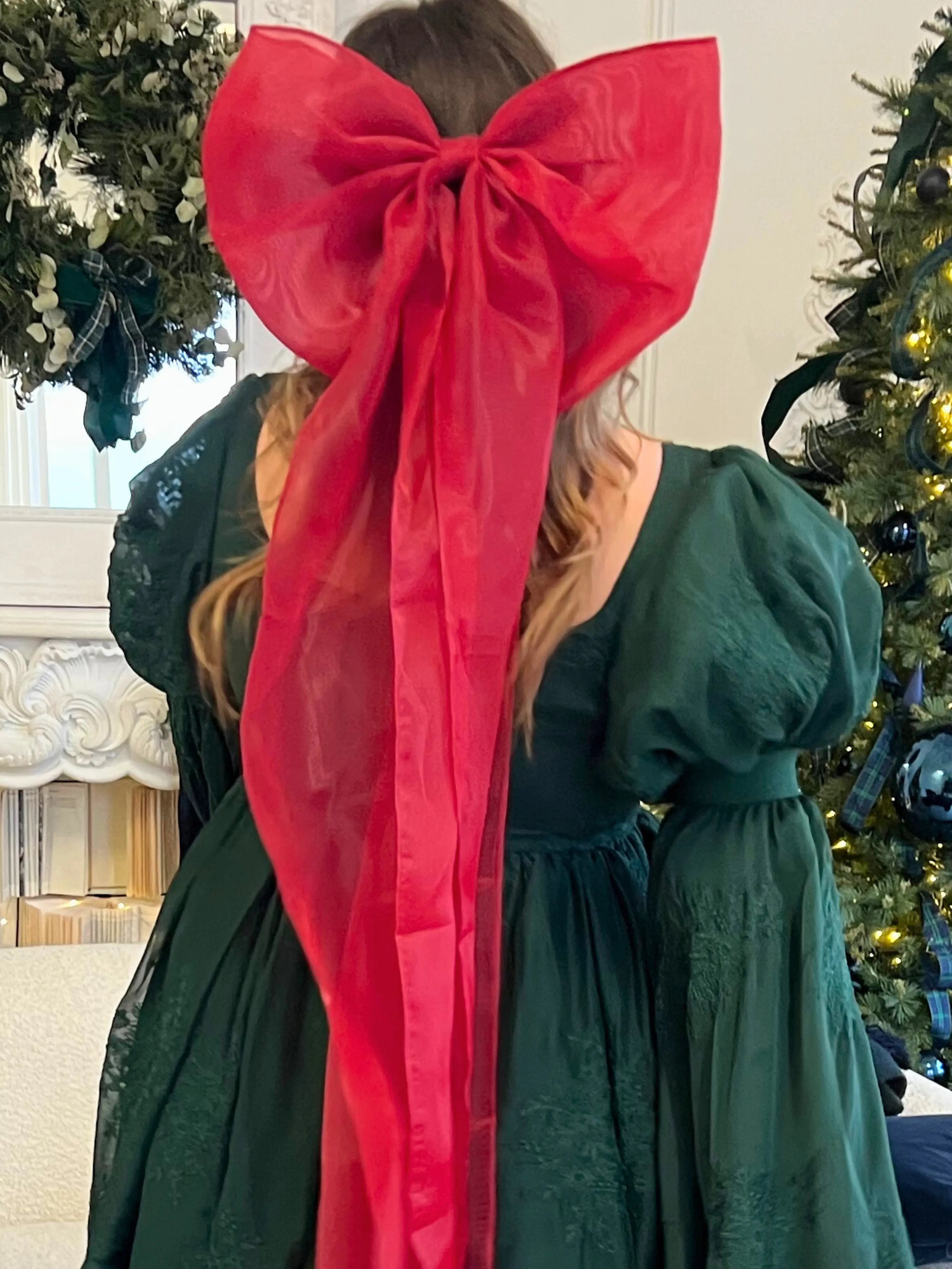 Bright red giant flutter bow on long a model with blonde hair wearing a green holiday mini dress by a Christmas tree.