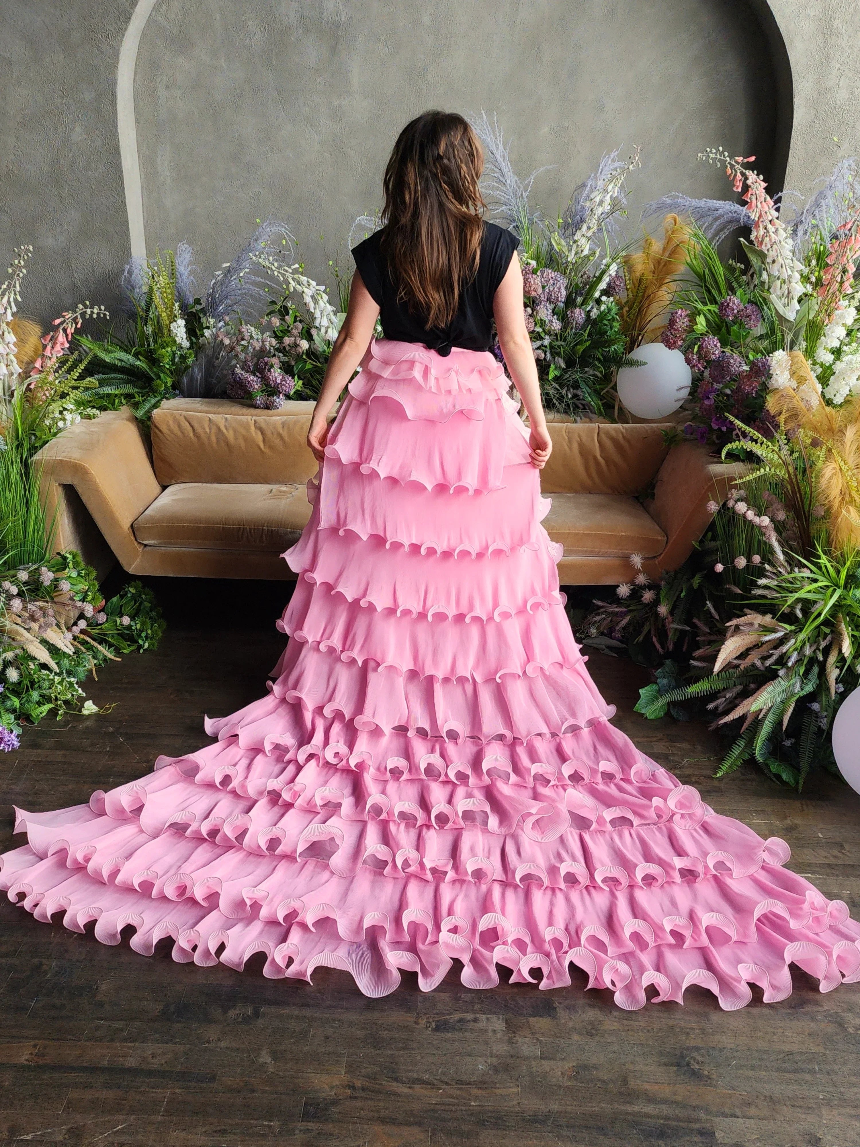 Voluminous pink Dahlia Ruffle Skirt with long train and snap closure buttons worn by woman surrounded by flowers for a fairytale scene.