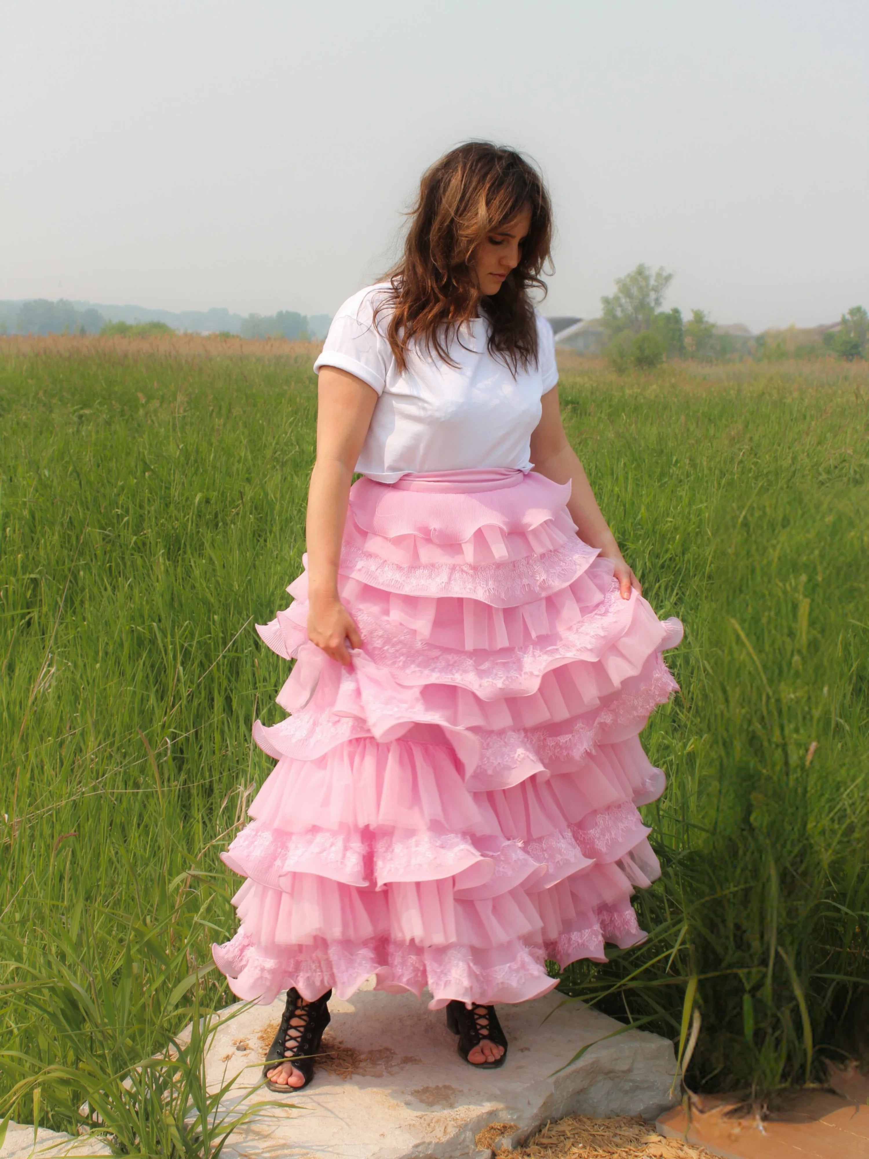 Pink multi-tiered Dahlia Ruffle Skirt with snap closure buttons paired with a white t-shirt, worn by a woman in a field for a fantasy look.