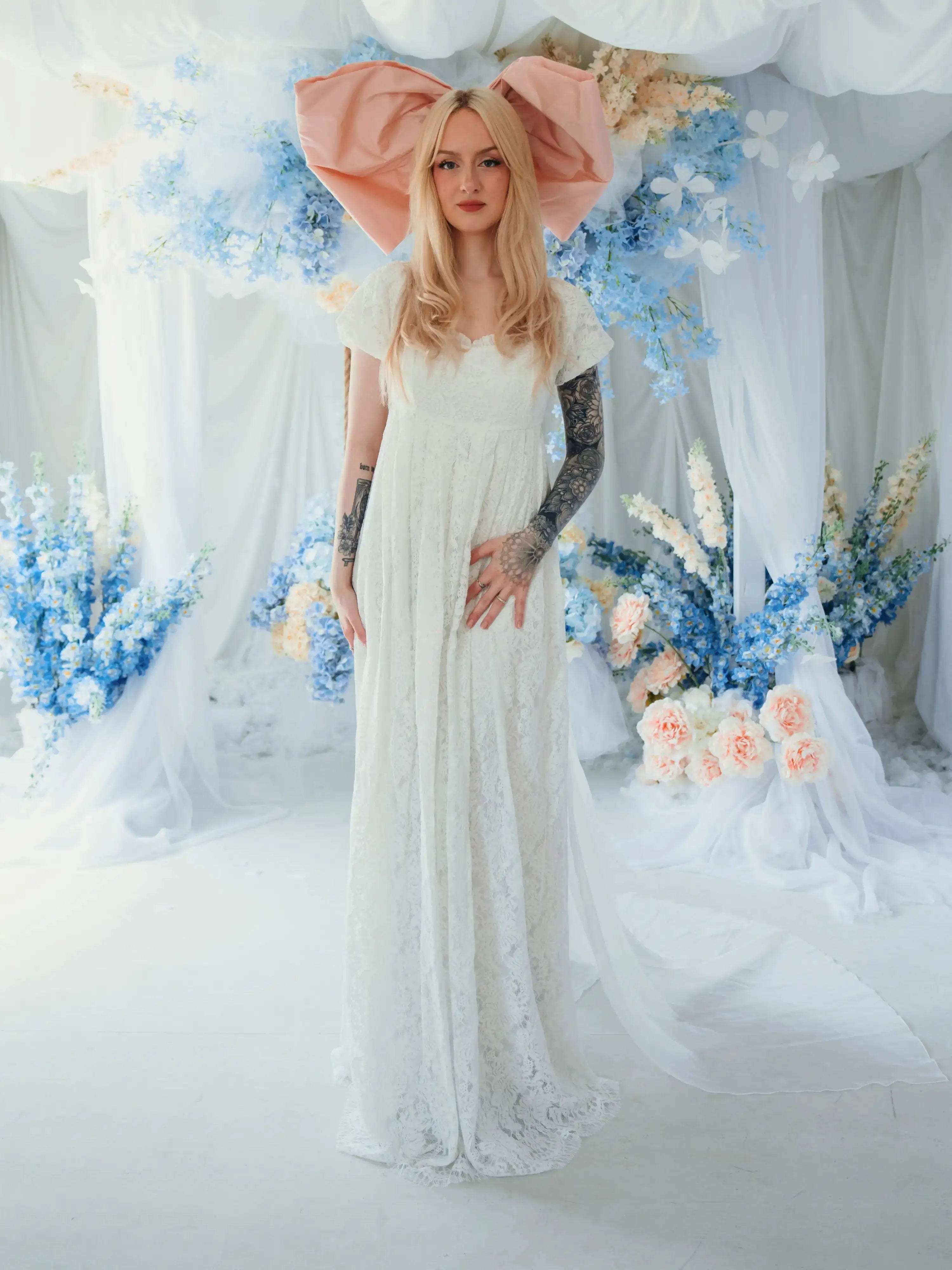 Dauphine Lace Gown bridal maxi dress with puff sleeves and empire waist, styled with oversized pink bow in a floral wedding setting.