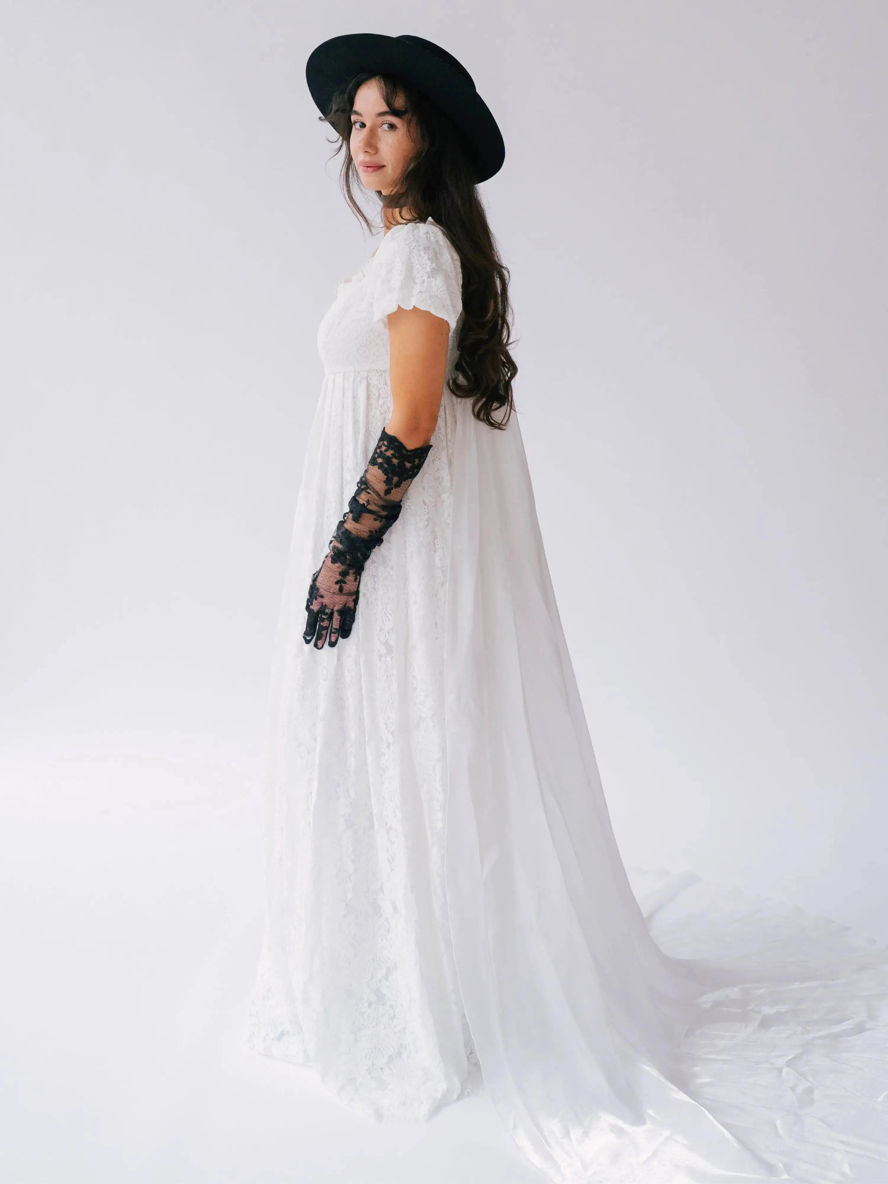 Side view of Dauphine Lace Gown with flowing train and puff sleeves, styled with black hat and lace gloves for a modern bridal look.
