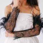 Delicate black lace arm sleeves with feather trim worn over a white ruffled mini dress.